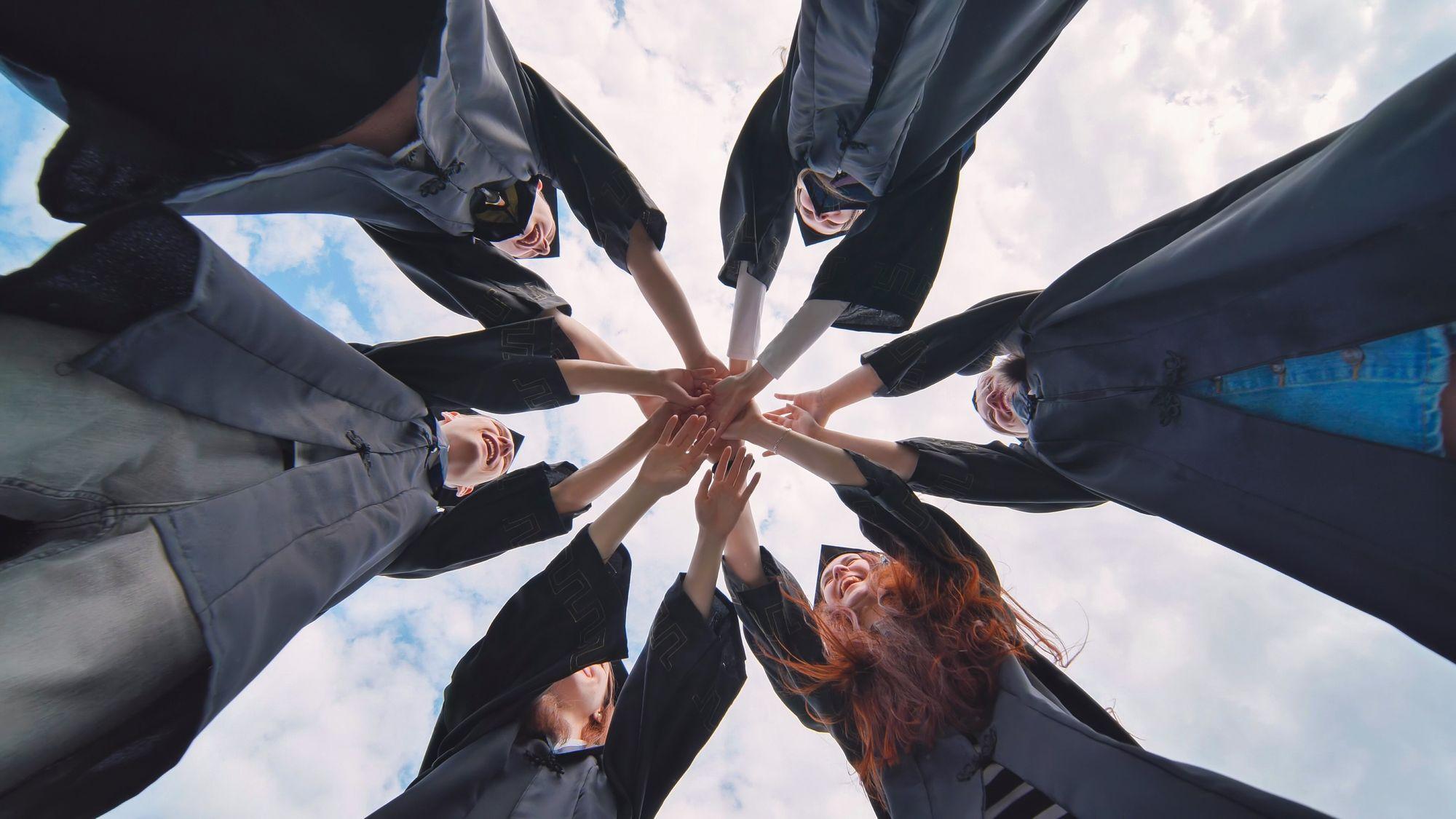 Graduates in caps and gowns joining their hands together in a circle against the sky.